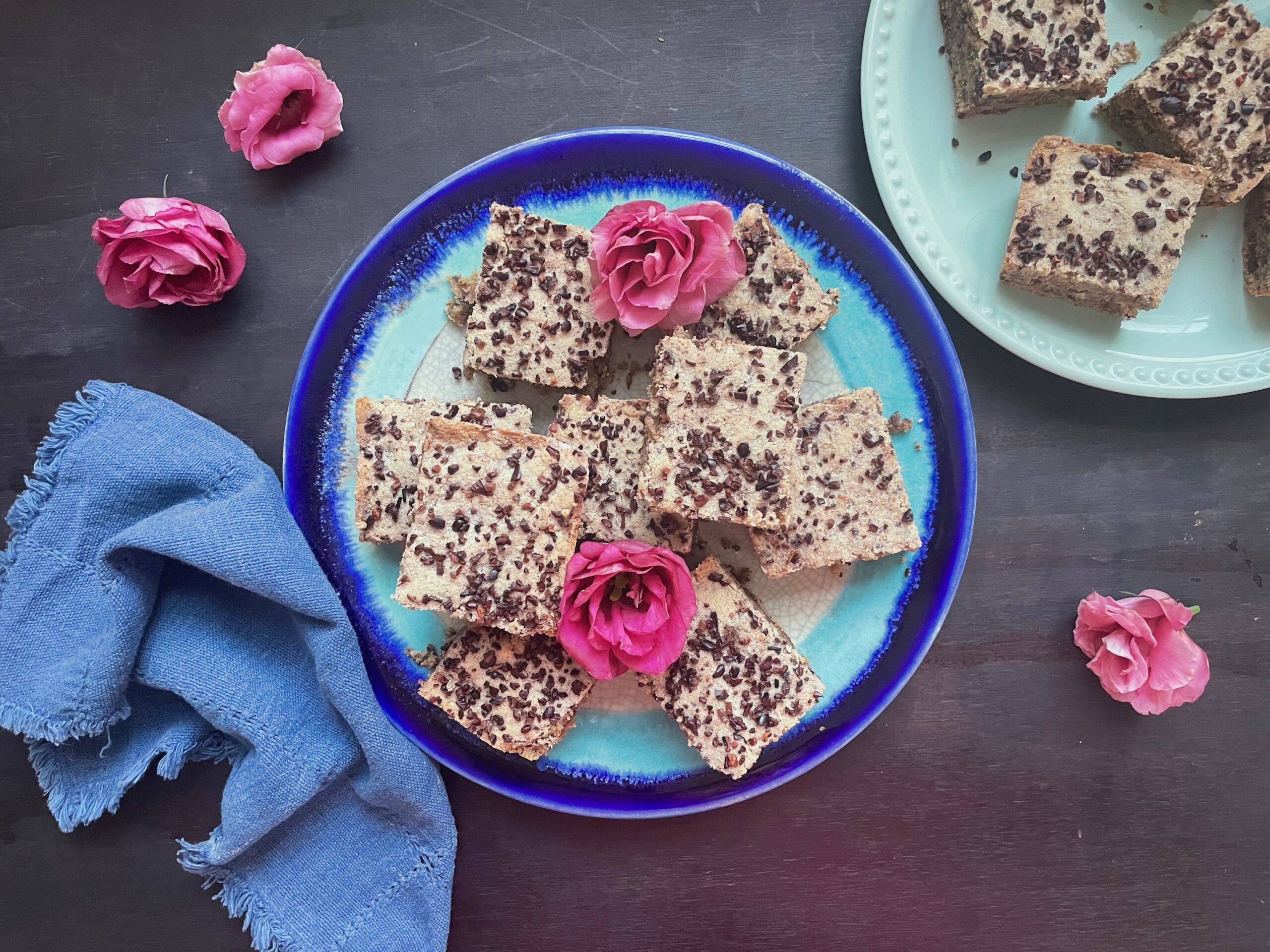 an angular arrangement of square bars sit atop a brilliant blue pottery plate. The cookie bars are mottled wiht cacao chips and two roses that are brilliant pink are nestled in the bars. two plates are whowen and the backround is black wood, a cotton neutral blue tea towel is the the left of one of the plants. the other plate is in the top right courner and you only see 1/3 of the plate.