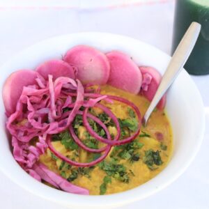 A bowl of cauliflower curry sits on a white background covered with pickeled daikon and cabbage...with a glass of spirulina pinapple juice in on a white background