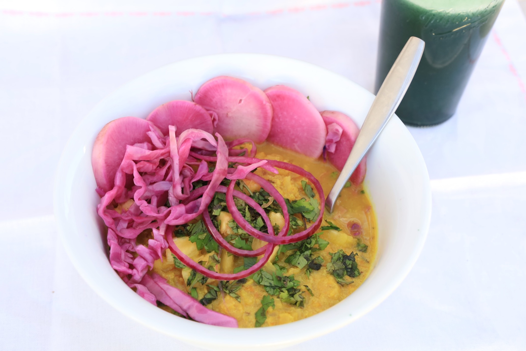 A bowl of cauliflower curry sits on a white background covered with pickeled daikon and cabbage...with a glass of spirulina pinapple juice in on a white background