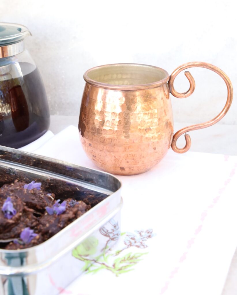 a stainless steel box of chocolates is featured from the side angle with the background containing a glass tea pot of dark red liquid, hibiscus tea next to a bright copper mug which is the centerpiece of the photo.