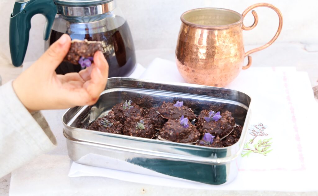 a stainless steel rectangular box hold around 21 mounds of chocolate truffles each topped with a purple lavender flower. A child's hand holds one truffle face up.