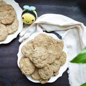 two white plates contain yummy cookies that are 3 inches in diameter. They are resting on a black stained table and a textile cotton cloth of natural fiber is draped around the plate to the position of 3 o clock. The other plate is at 12 o clocK. A stuffed bumble bee is at 1 o clock. A few plant leaves have made their way into the picture