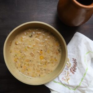 A light tan pottery bowl contains a soup that is similar in color to the bowl and is flecked with small rounds of carrot and squares of cabbage. A white linen napkin embrowderied wiht bluebonnets is on the side and a tall clay drinking vessel is shown at the top right corner.