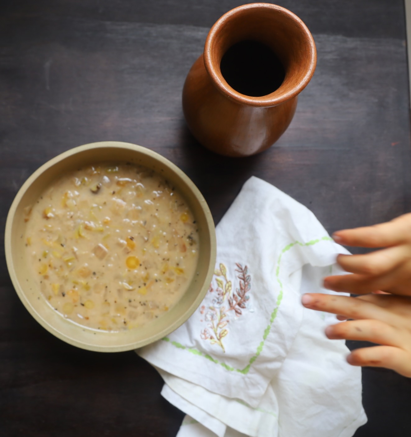 A light tan pottery bowl contains a soup that is similar in color to the bowl and is flecked with small rounds of carrot and squares of cabbage. A white linen napkin embrowderied wiht bluebonnets is on the side and a tall clay drinking vessel is shown at the top right corner. A childs hands are reaching for the bowl above the napkin. The photo is in color. The child's hands are small.