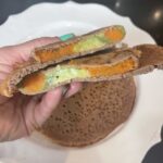 a brown crepe cut in half to show the filling of sweet potato and avocado in strips. A turquose manicured hand is used to hold the crep in front of a white platter that holds other crepes.
