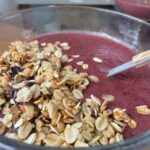 a clear smoothie bowl is filled with a smoothie with a half moon covering of date granola
