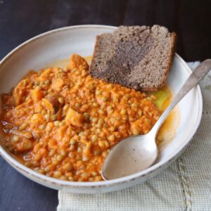 An off white pottery dish with a 1 in lip holds a large mound of lentil stew. The place also has Brown Irish Soda Bread and a large sliver spoon. Underneath is an off white linen napkin.