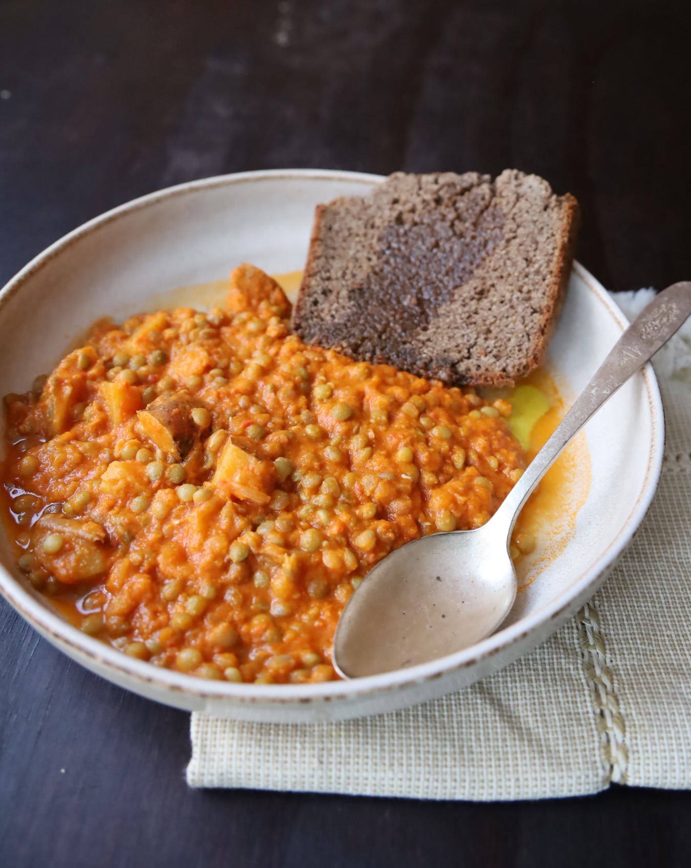 An off white pottery dish with a 1 in lip holds a large mound of lentil stew. The place also has Brown Irish Soda Bread and a large sliver spoon. Underneath is an off white linen napkin.