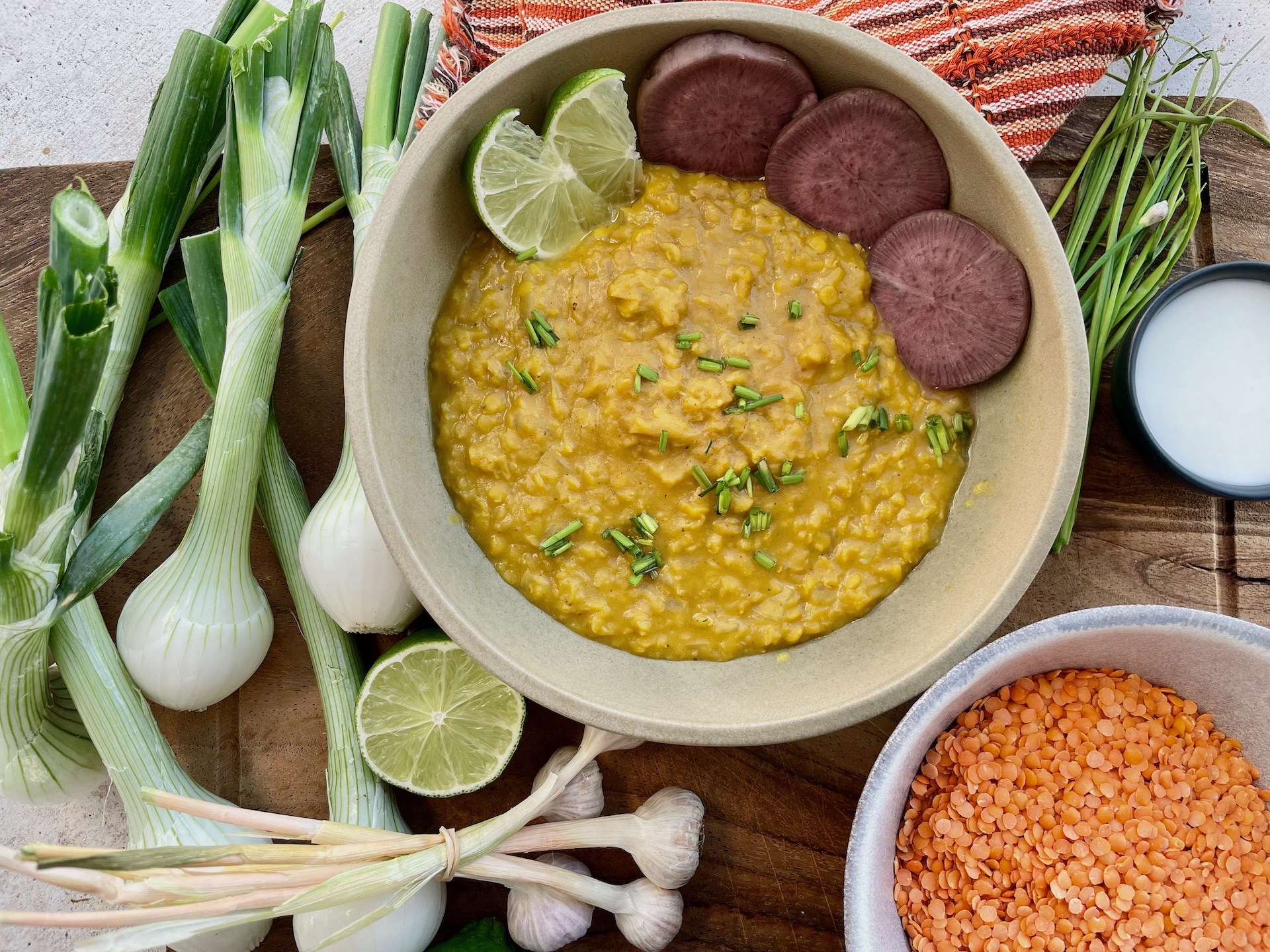 a bowl of lentils is cooked and plated with pickled daikon radish. Surrounded by green onions, garlic bunches, dry lentils, and a yogurt dill sauce.