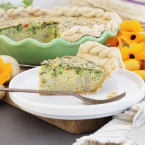 A vegetable quiche in a green pottery pie dish is surrounded by orange flours, bright red peppers, and potatoes. A slice is cut out of the whole pie and put on the side. This photo is a side view of the slice.