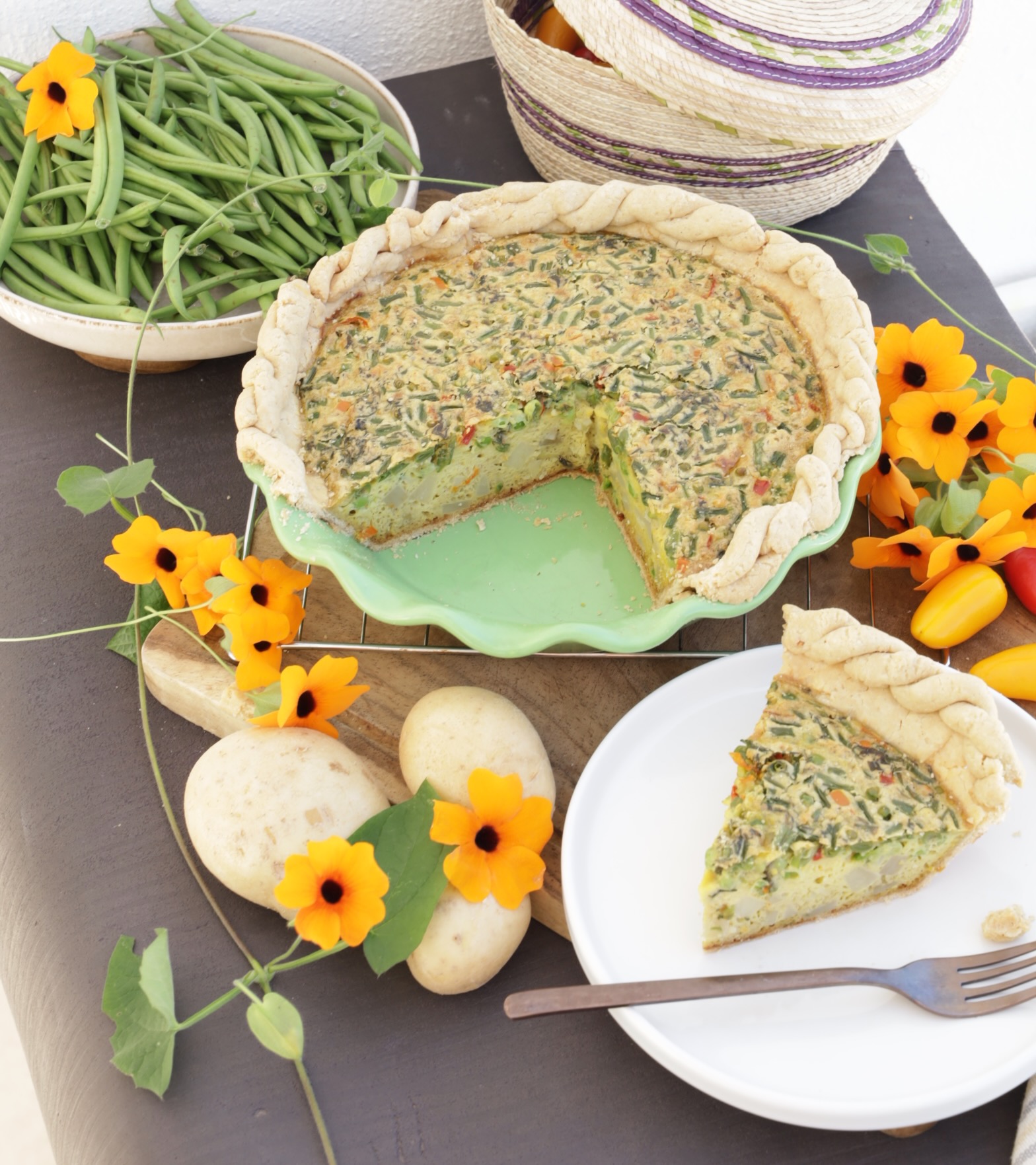 A vegetable quiche in a green pottery pie dish is surrounded by orange flours, bright red peppers, and potatoes. A slice is cut out of the whole pie and put on the side.