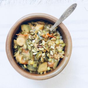 a pottery bowl is filled with summer stew. A silver spoon is plunged in the bowl and it is topped with toasted pumpkin seeds.