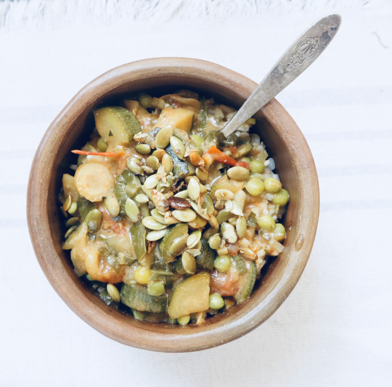 a pottery bowl is filled with summer stew. A silver spoon is plunged in the bowl and it is topped with toasted pumpkin seeds.