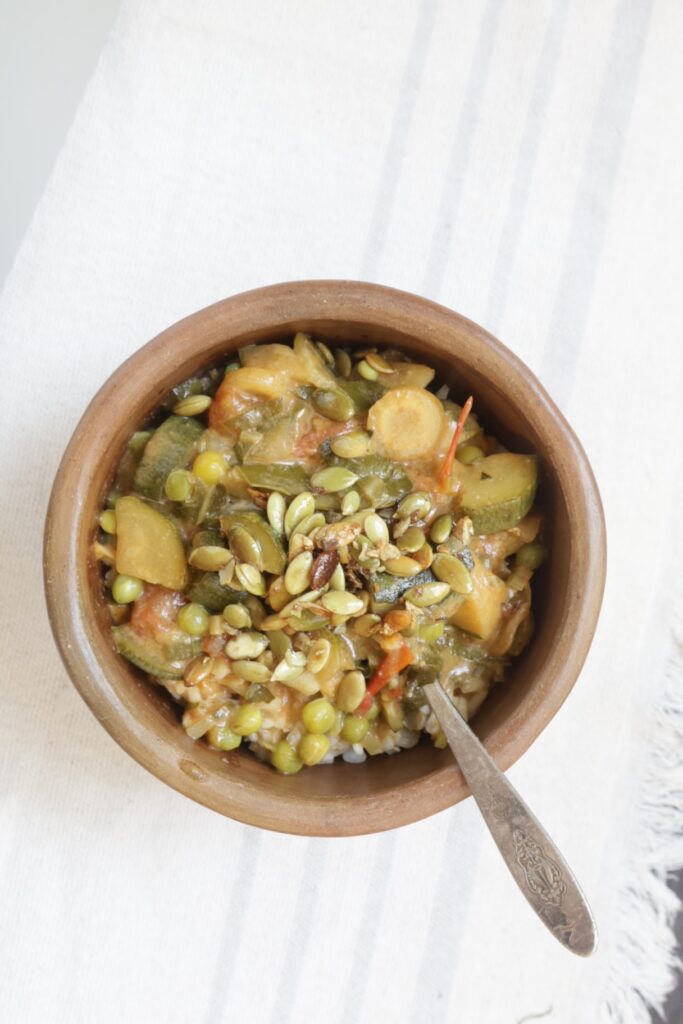 a pottery bowl is filled with summer stew. A silver spoon is plunged in the bowl and it is topped with toasted pumpkin seeds.