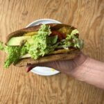 A polished hand holds a wrap filled with egg, avocado, lettuce, tomato, and sunflower seeds