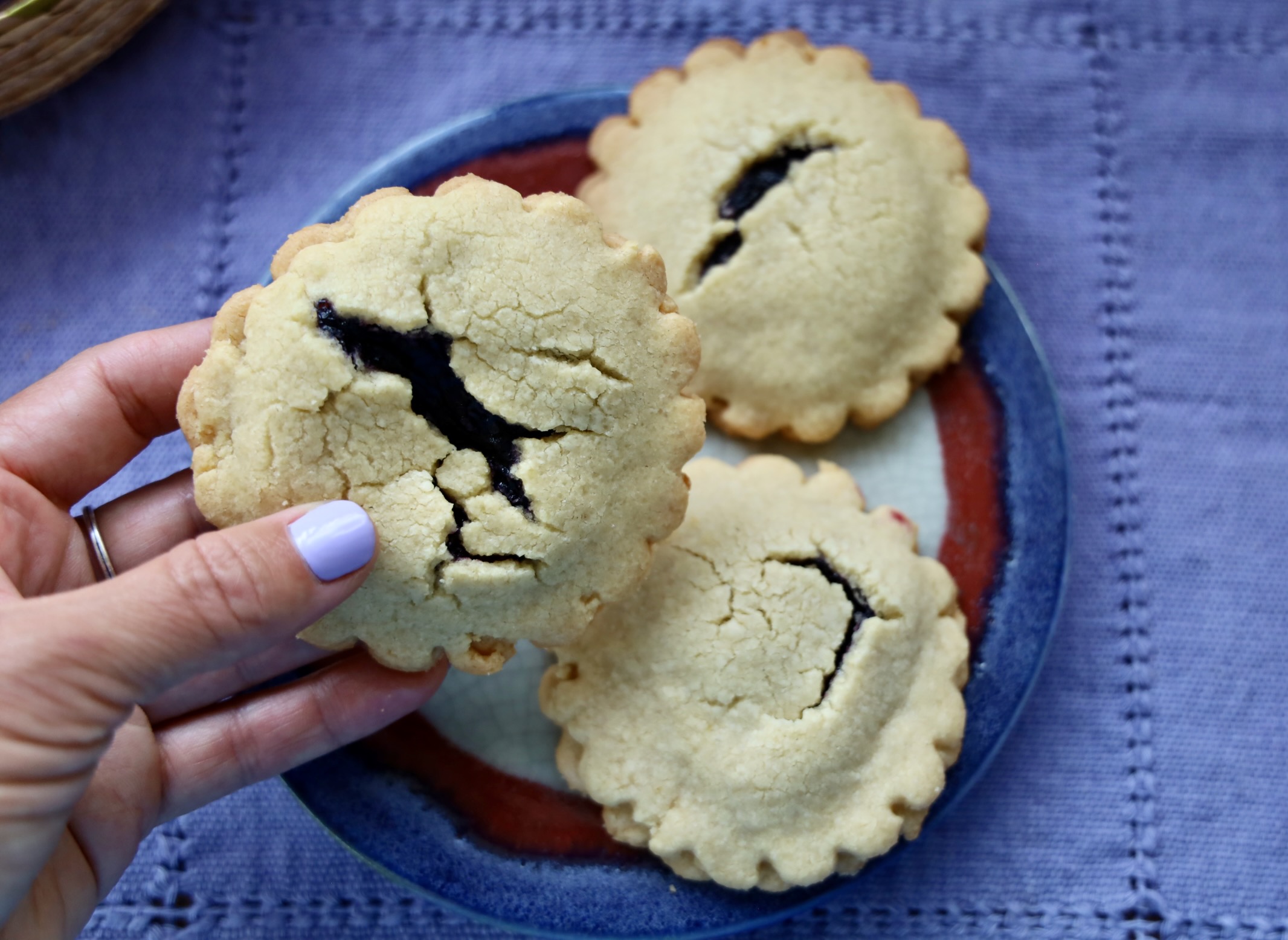 A purple polished hand holds a jam filled gluten free cookie with the other cookies in the background on top of a purple linen tea towel.