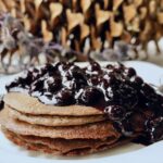 A stack of Blender Pancakes is covered with a large amount of Blueberry Jam Compote on a white plate. The angle is from the side of the pancakes that are on a white plate. A pinecone is out of focus in the background.