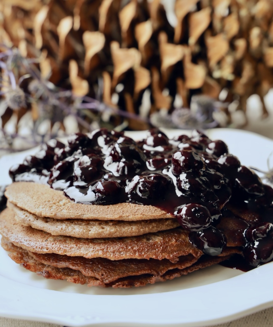 A stack of Blender Pancakes is covered with a large amount of Blueberry Jam Compote on a white plate. The angle is from the side of the pancakes that are on a white plate. A pinecone is out of focus in the background.
