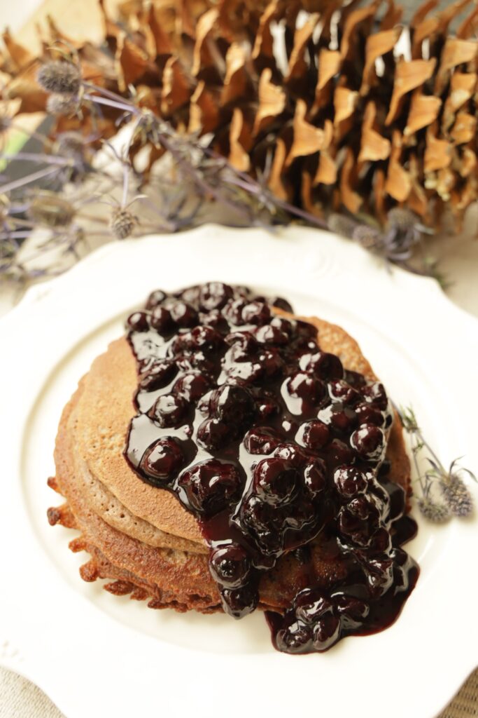 A stack of Blender Pancakes is covered with a large amount of Blueberry Jam Compote on a white plate. The angle is from the top of the pancakes that are on a white plate. A pinecone and purple thistles are out of focus in the background.