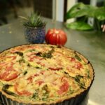 A tomato basil quiche is presented in a tart pan. Tomatoes and basils burst from top of the egg and cheese crust. The pie rests on a stainless steel table with a table and plants in the background.