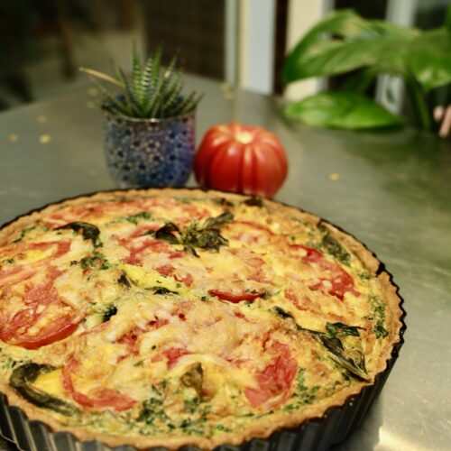 A tomato basil quiche is presented in a tart pan. Tomatoes and basils burst from top of the egg and cheese crust. The pie rests on a stainless steel table with a table and plants in the background.