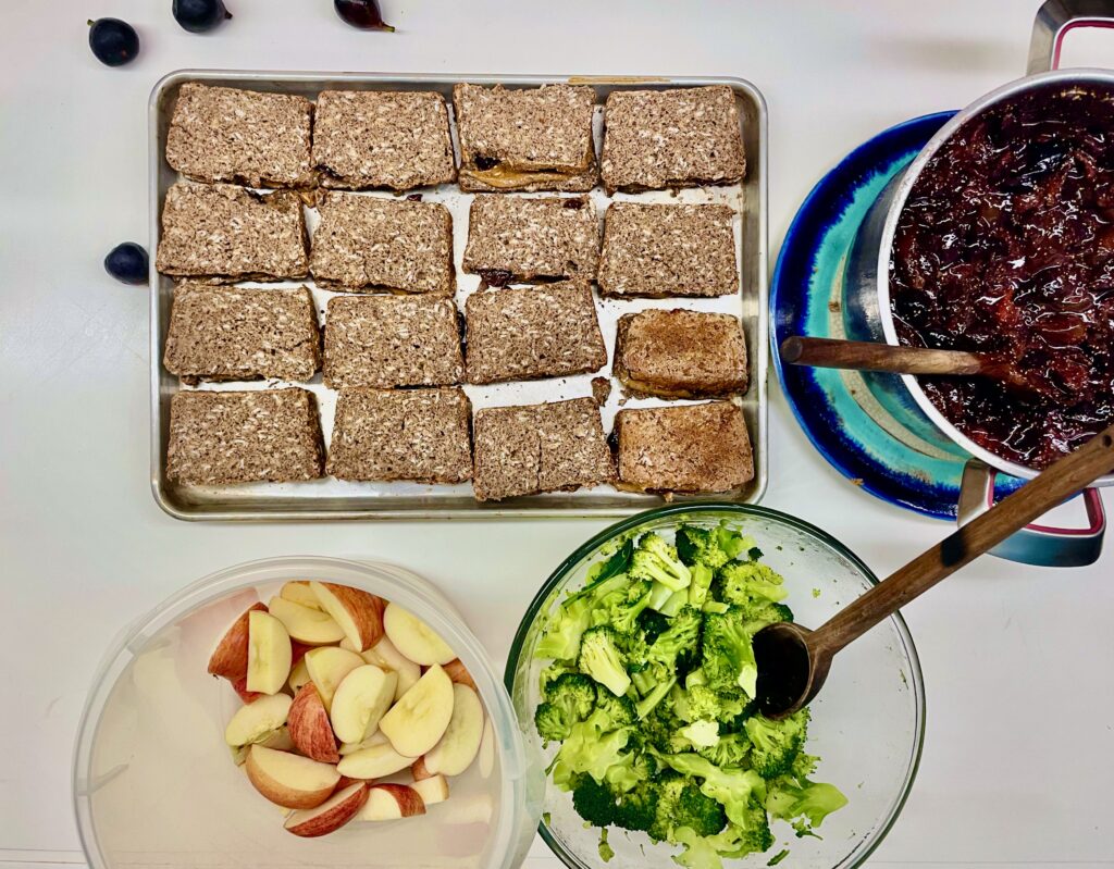 A cookie sheet has a tray of 16 nut butter and fig sandwhiches and a bowl of broccoli and bowl of apples.